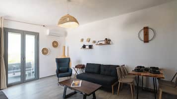 A modern living room with a sofa, chairs, a coffee table, and a dining table. There is a large window with curtains, a hanging light fixture, and wall decorations.