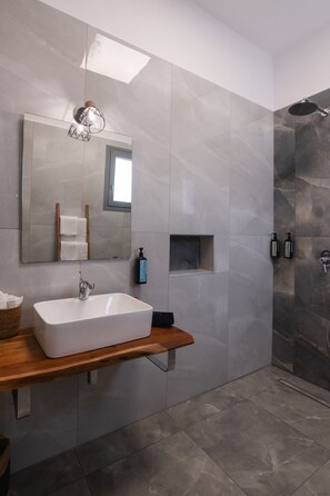 A modern bathroom with a rectangular sink, a mirror, a shower, and a small shelf with toiletries. - Filia Skyros Suites (Skiros)
