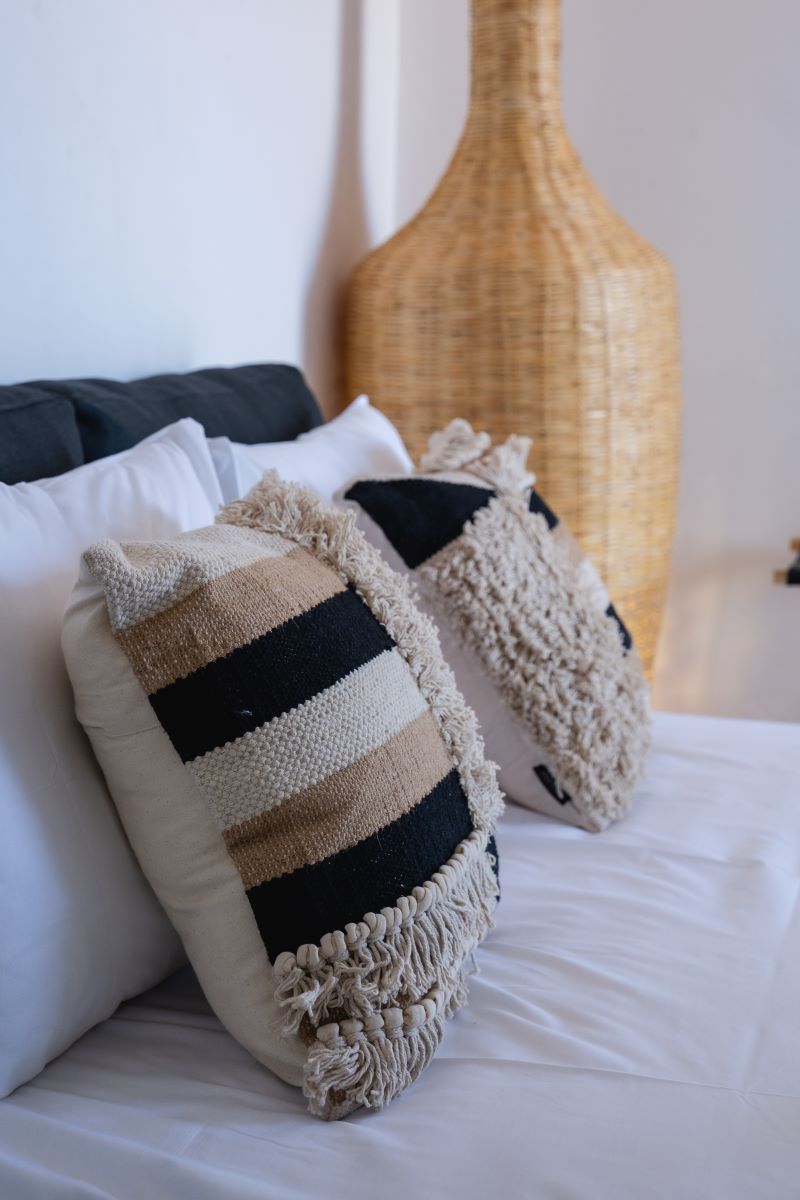 Una cama con dos almohadones decorativos, un jarrón tejido y una pared lisa.