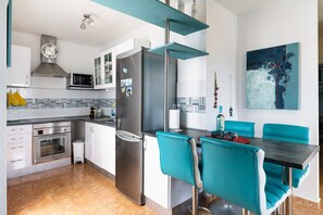 Una cocina moderna con electrodomésticos de acero inoxidable, una zona de comedor con sillas color turquesa y un cuadro abstracto azul en la pared.