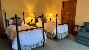 Una habitación con dos camas con dosel, cada una con una mesita de noche y una lámpara. Hay una puerta de madera y una silla azul en la habitación.