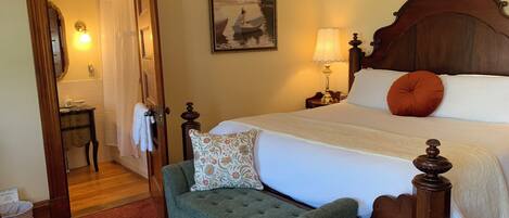 A neatly arranged bedroom with a wooden bed, a green armchair, a nightstand with a lamp, and a painting on the wall.