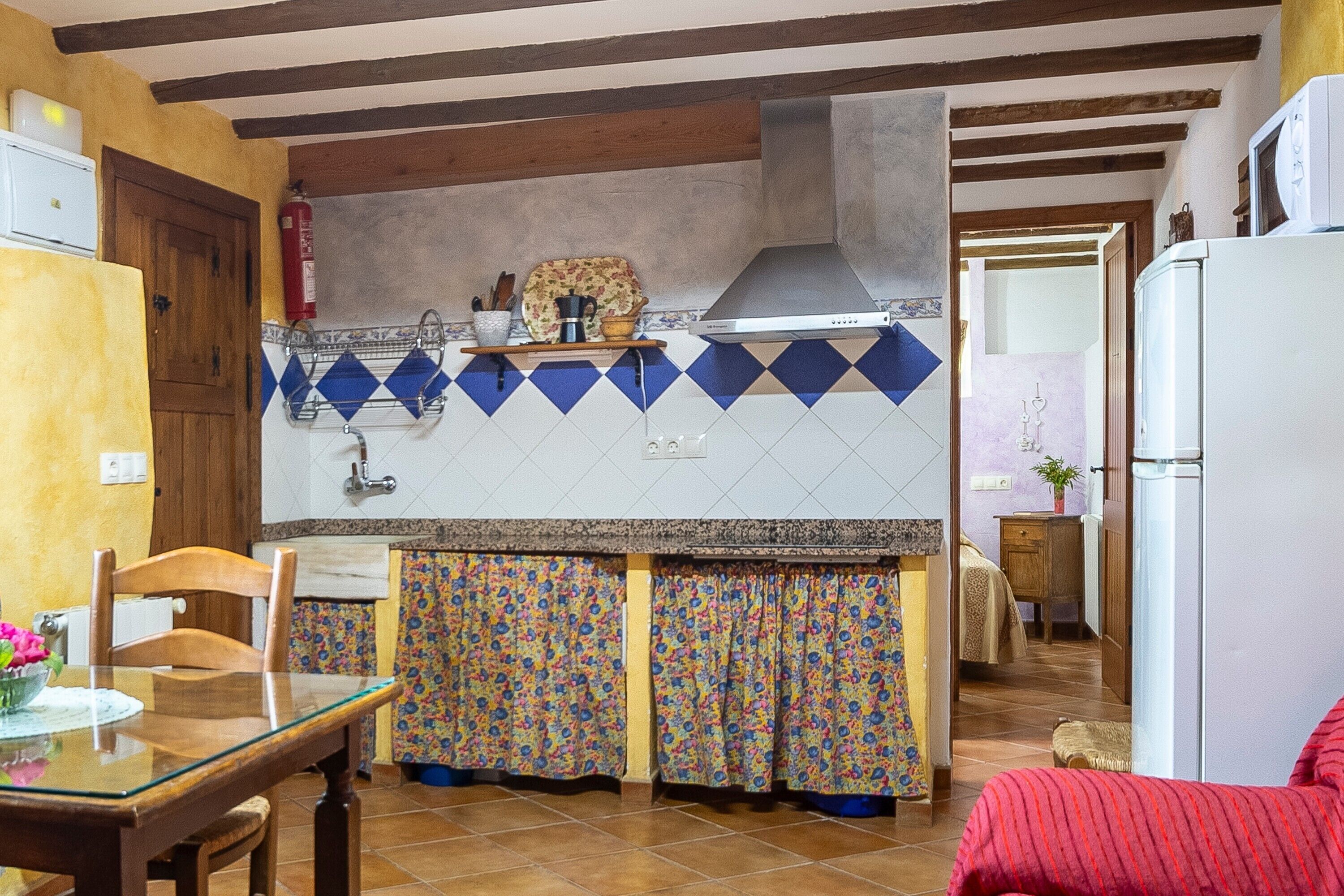Cozinha com painel de azulejos, vigas de madeira, geladeira e área de jantar com mesa e cadeiras.