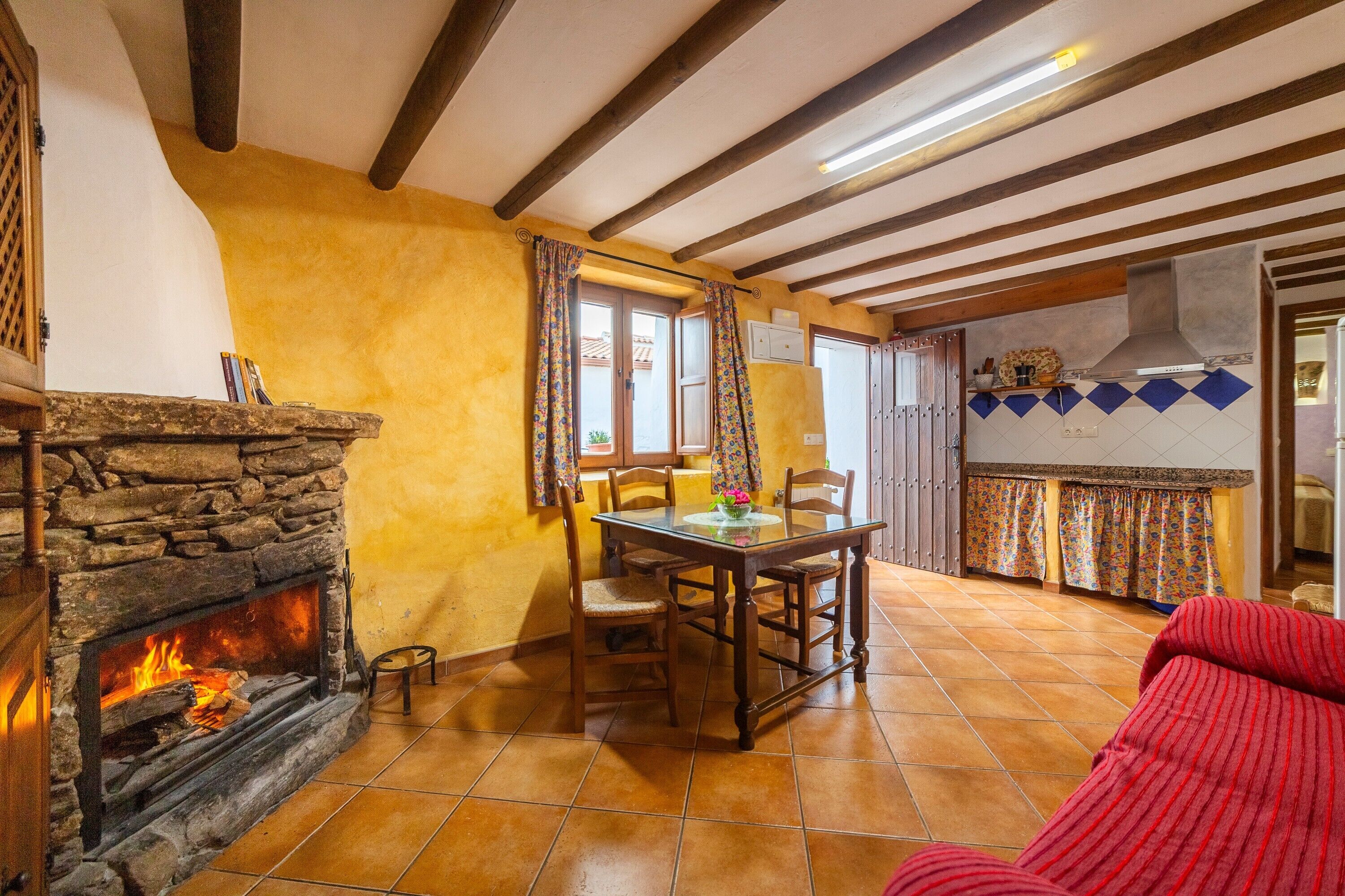 Una habitación acogedora con chimenea de piedra, vigas de madera, una mesa de comedor con sillas y un sofá rojo.