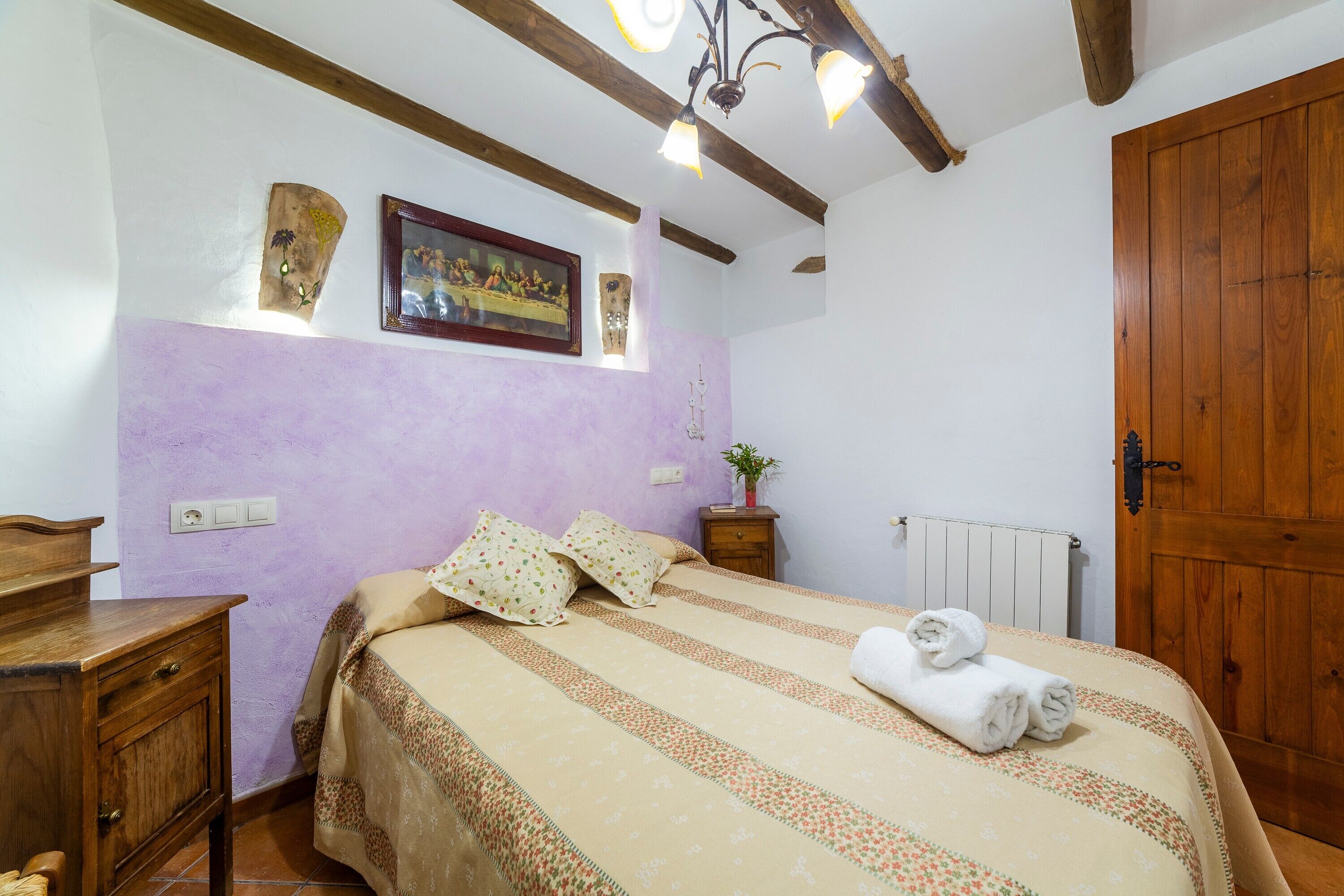 Un dormitorio con cama, cabecero de madera, armario de madera, mesita de noche, estante en la pared, un cuadro enmarcado y una lámpara de techo.