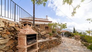 Zona de cuina exterior de pedra amb forn de pedra, un mur de pedra i una vista d'una casa al fons.