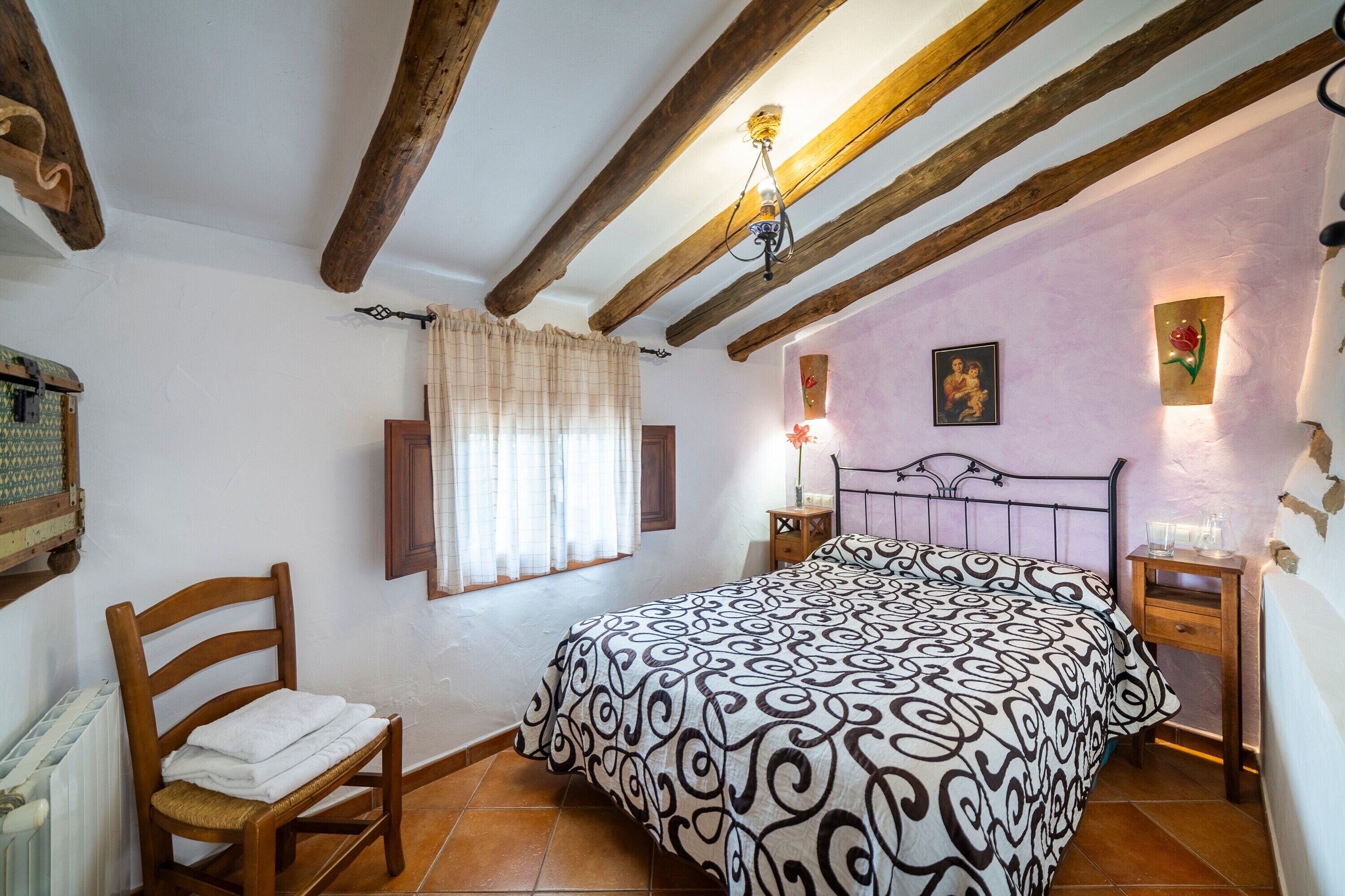 A bedroom with a bed, wooden chair, nightstands, and a ceiling with exposed beams.