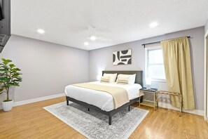 Une chambre à coucher avec un lit, une table de chevet, un ventilateur de plafond, une plante en pot et une fenêtre avec des rideaux.