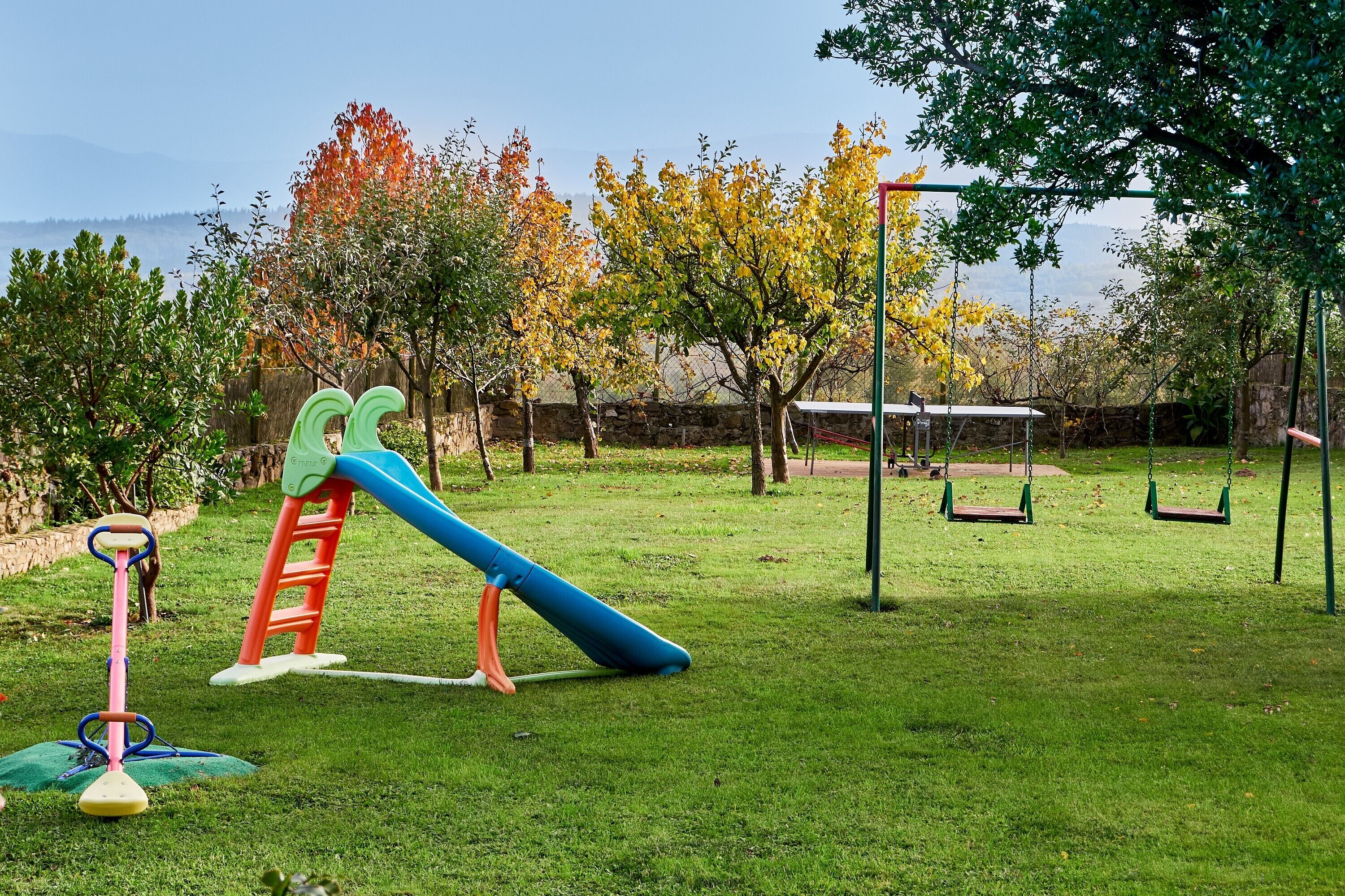 Un parque infantil con columpios y un tobogán, rodeado de árboles y un muro de piedra.
