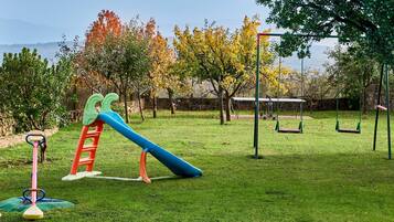 A playground with swings and a slide, surrounded by trees and a stone wall.