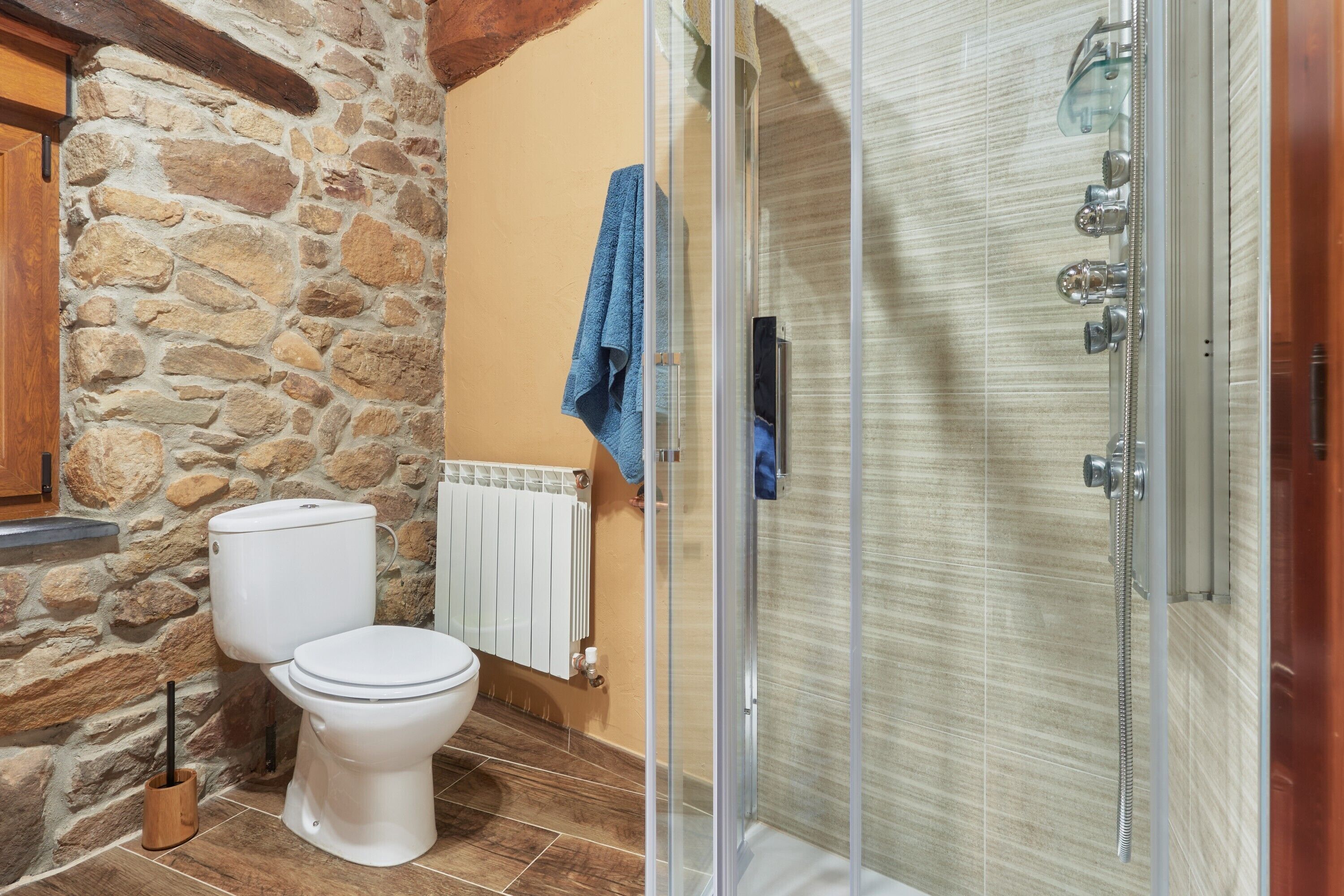 Une salle de bain avec un mur en pierre, des toilettes blanches, une douche en verre et une serviette bleue accrochée au mur.