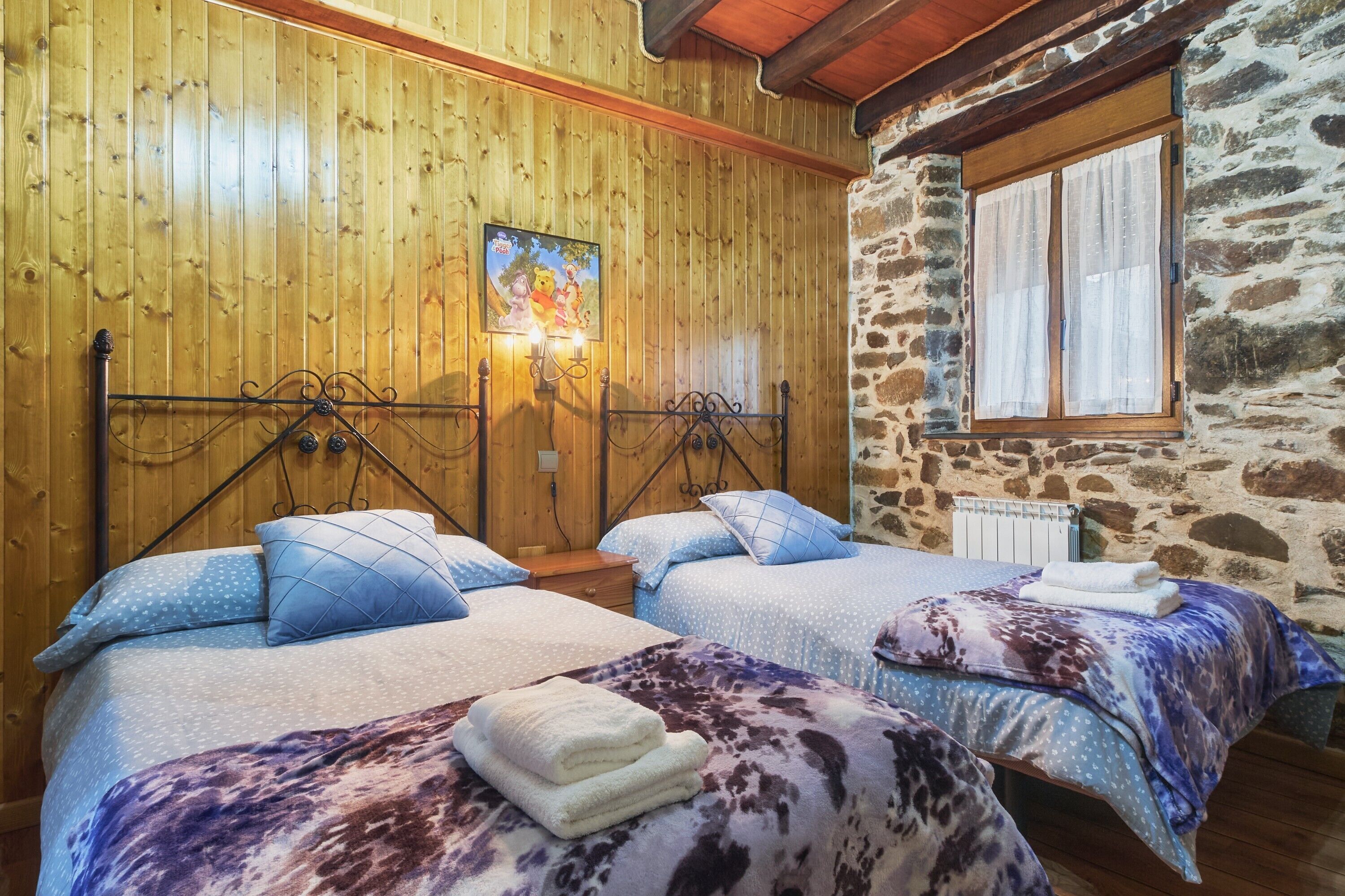 A room with two beds, wooden walls, a stone wall, a window with curtains, and a picture on the wall.