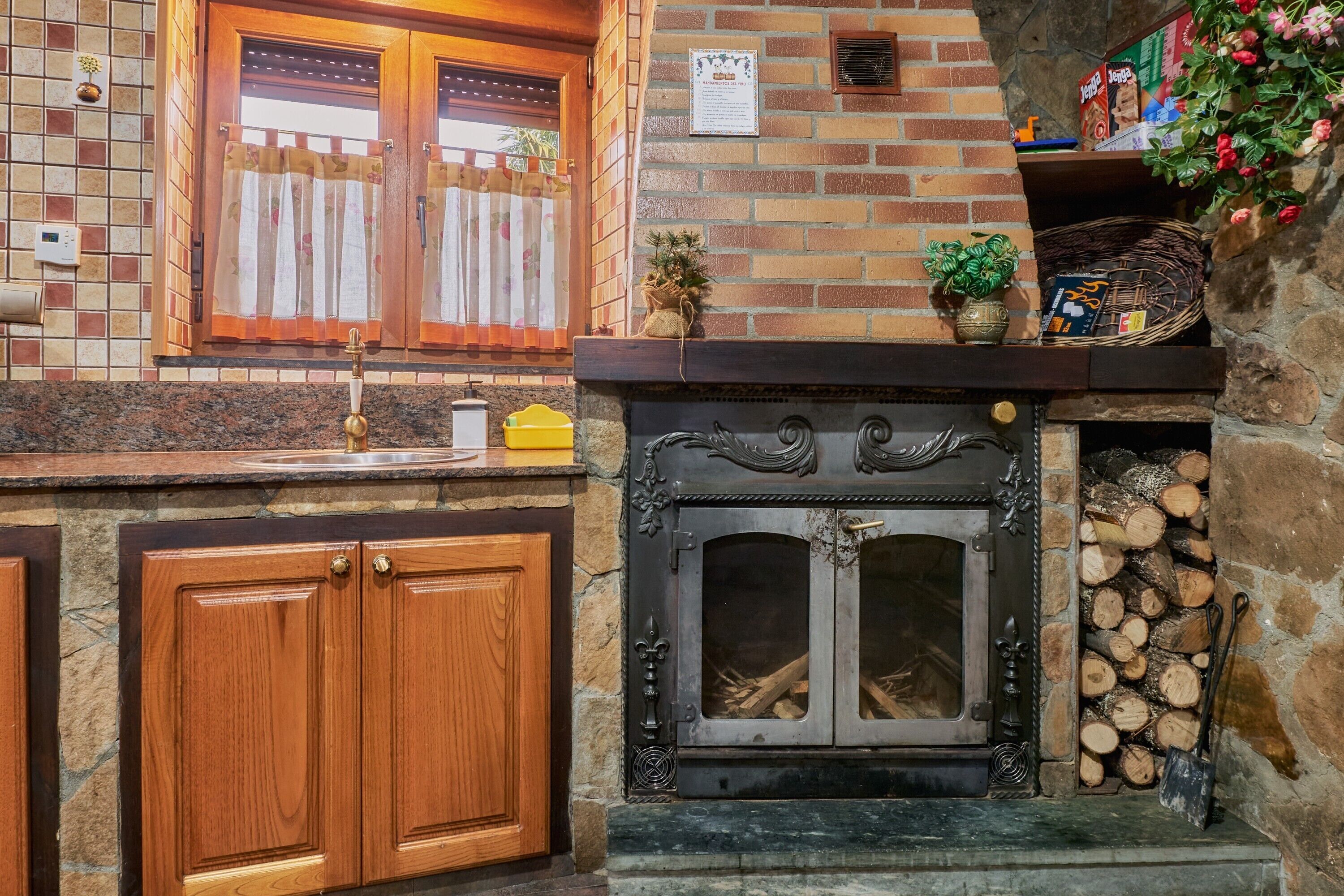 Una cocina con encimera de piedra, armarios de madera, chimenea con leña y una ventana con cortinas.
