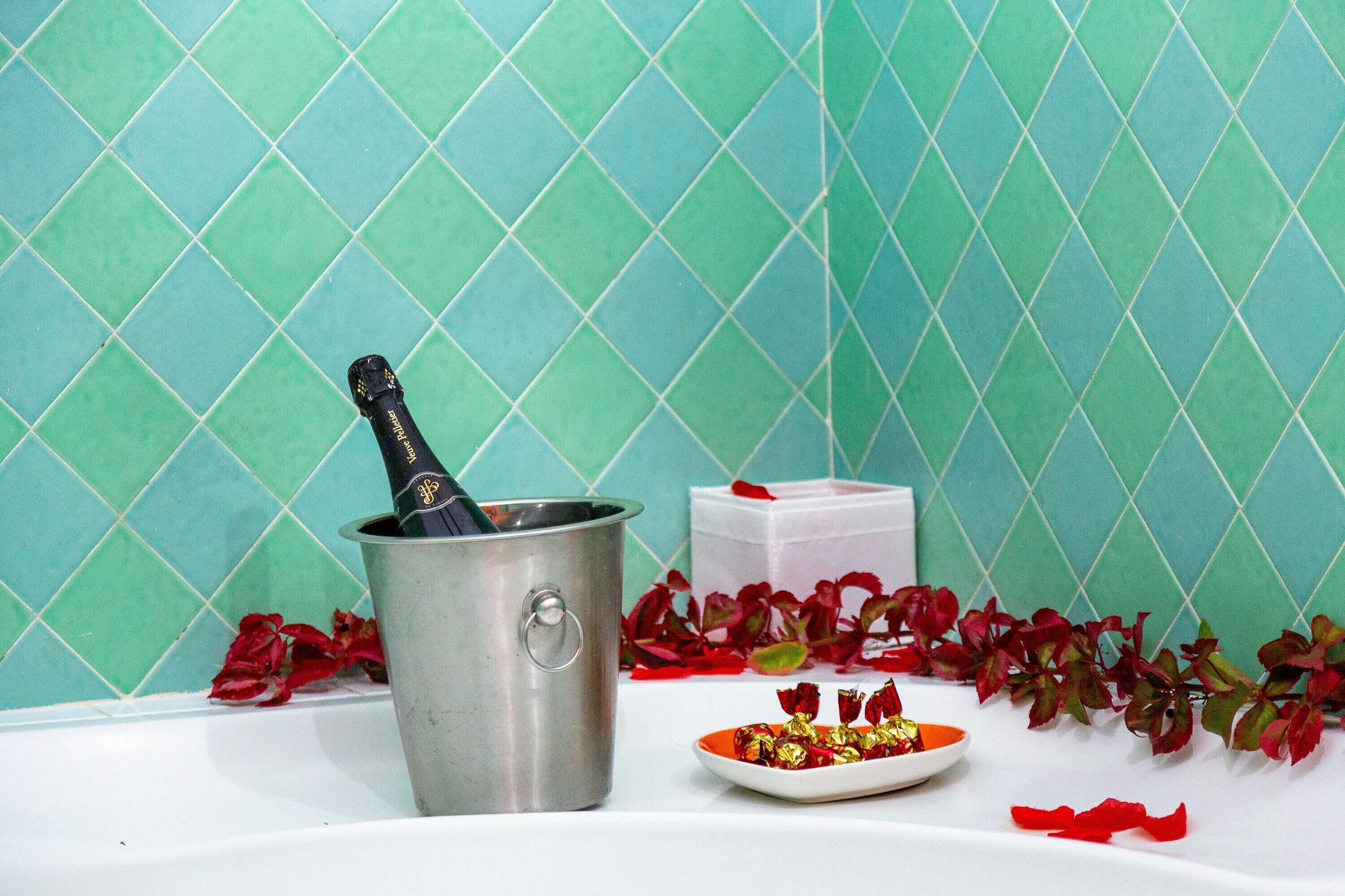 Une salle de bain avec un mur recouvert de carreaux verts, un seau métallique, une boîte cadeau blanche et une assiette avec des chocolats emballés.
