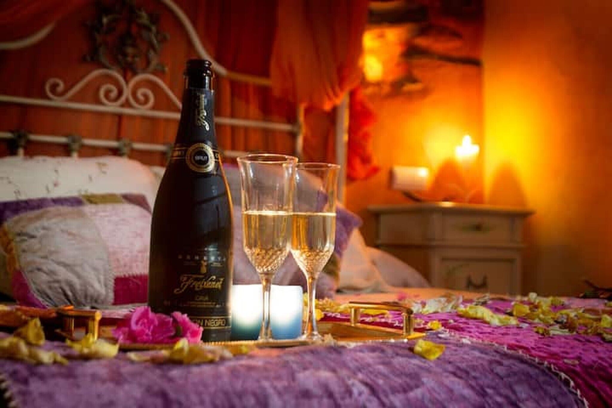 Champagnefles, twee glaasjes en een kaars op een bed met een paarse deken.