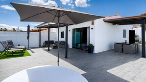 Patio esterno con ombrellone bianco, zona salotto e angolo cottura.