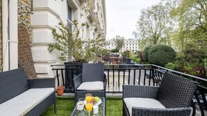 A balcony with wicker furniture, a glass table with fruit and drinks, and a view of a cityscape. - West Oasis - Escape From Chaos (London)