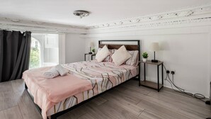 A neatly made bed with a pink blanket and pillows, a bedside table with a lamp, and a potted plant. - West Oasis - Escape From Chaos (London)