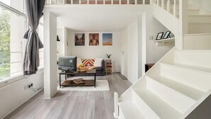 A modern living room with a sofa, coffee table, and a TV. A staircase leads to an upper level. - West Oasis - Escape From Chaos (London)