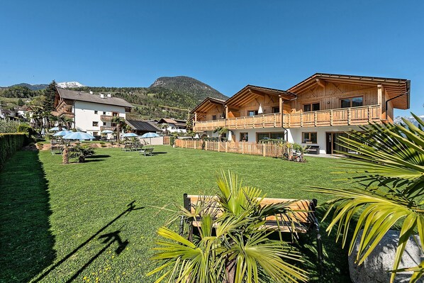 A two-story wooden chalet with a balcony, surrounded by a well-maintained lawn and a mountain backdrop.