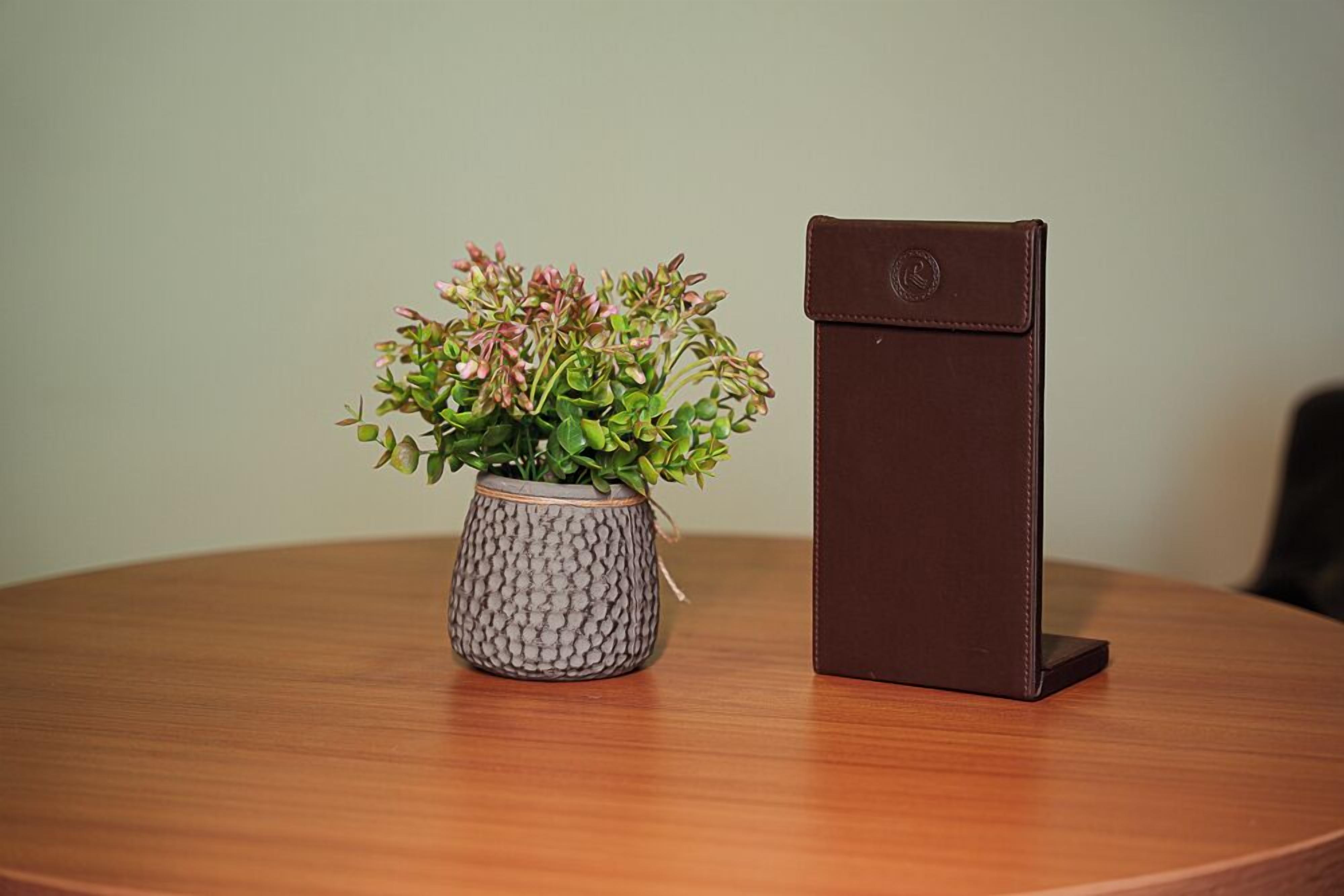A round wooden table with a textured grey vase containing green plants and a brown leather wallet.