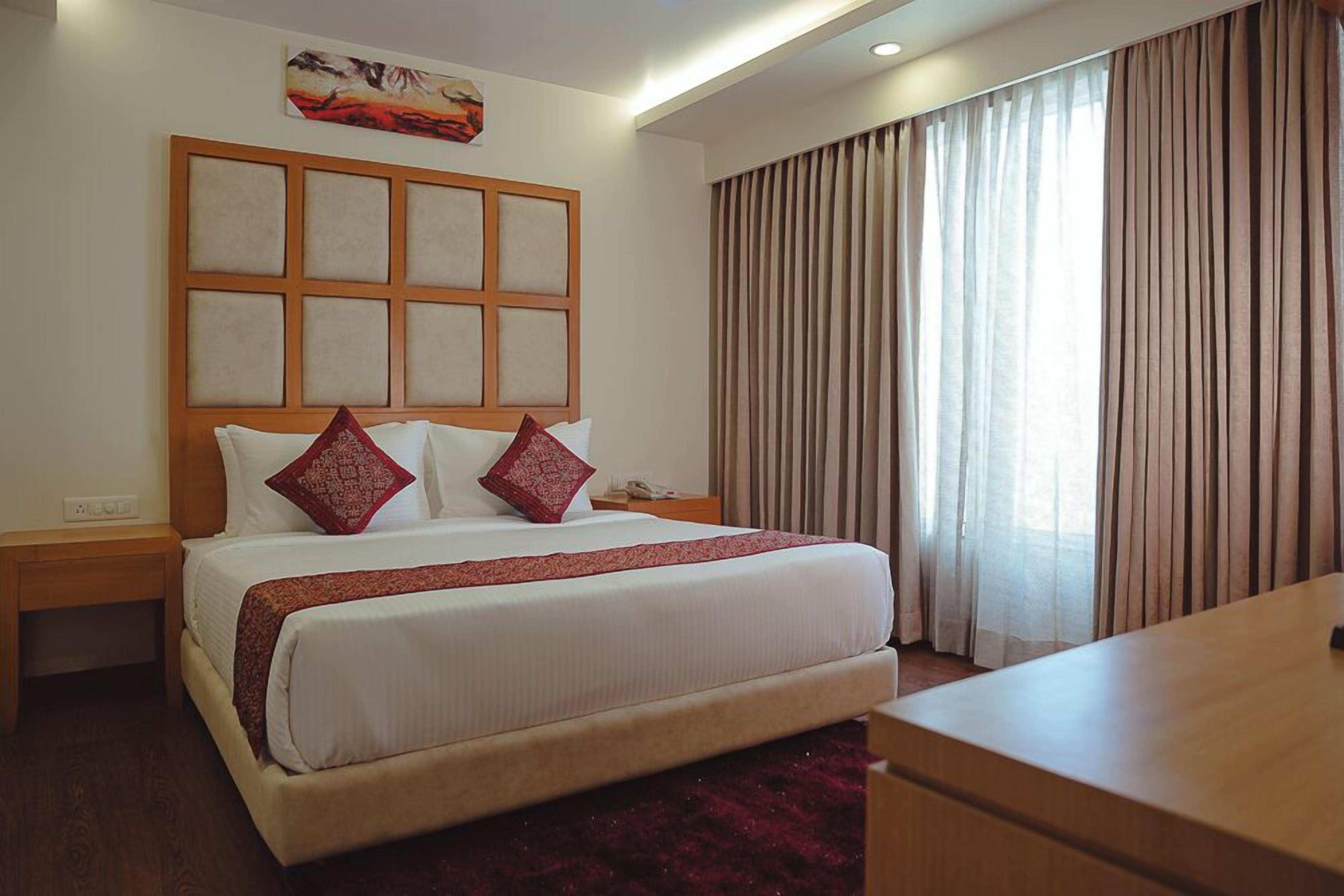 A hotel room with a large bed, a wooden headboard, a bedside table, a desk, and a window with curtains.