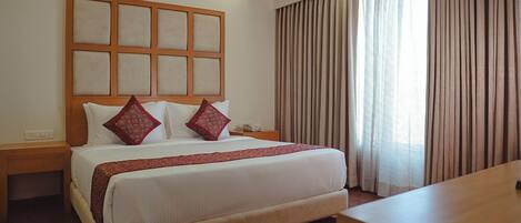 A hotel room with a large bed, a wooden headboard, a bedside table, a desk, and a window with curtains.
