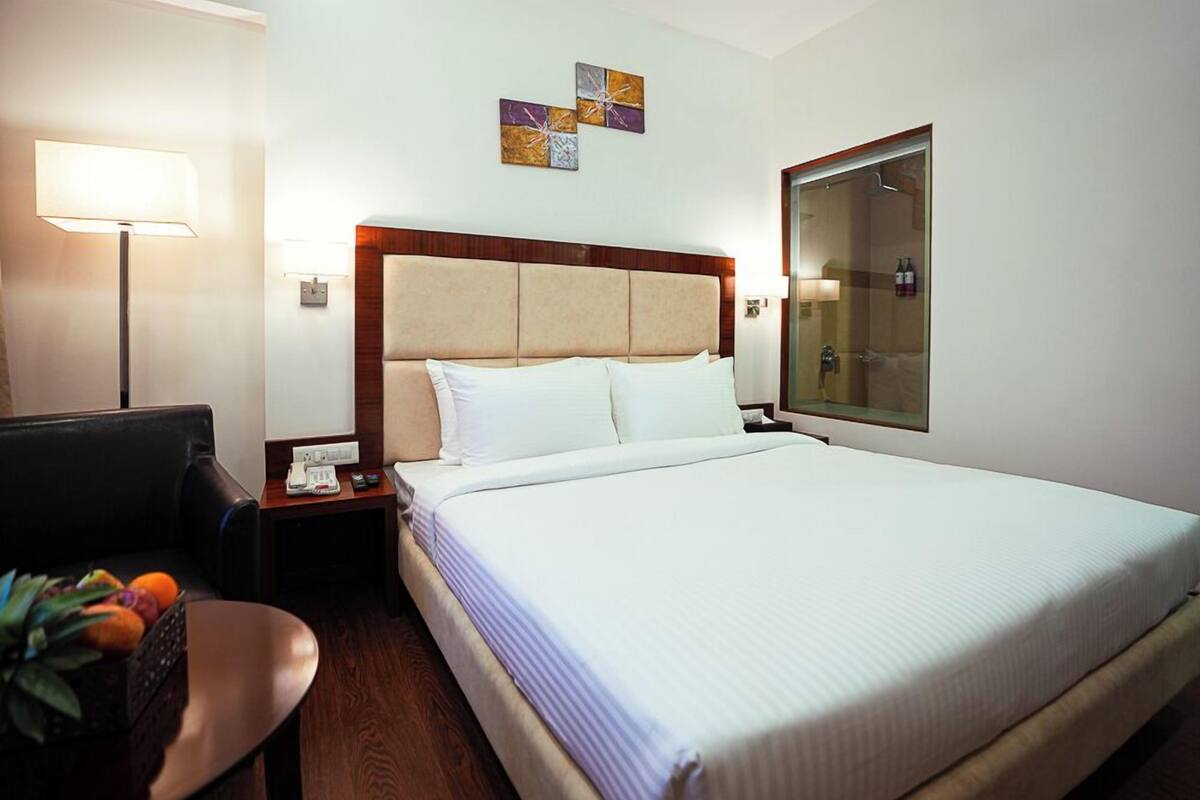 A hotel room with a bed, a desk with a lamp, a chair, and a small table with fruits.