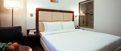 A hotel room with a bed, a desk with a lamp, a chair, and a small table with fruits.