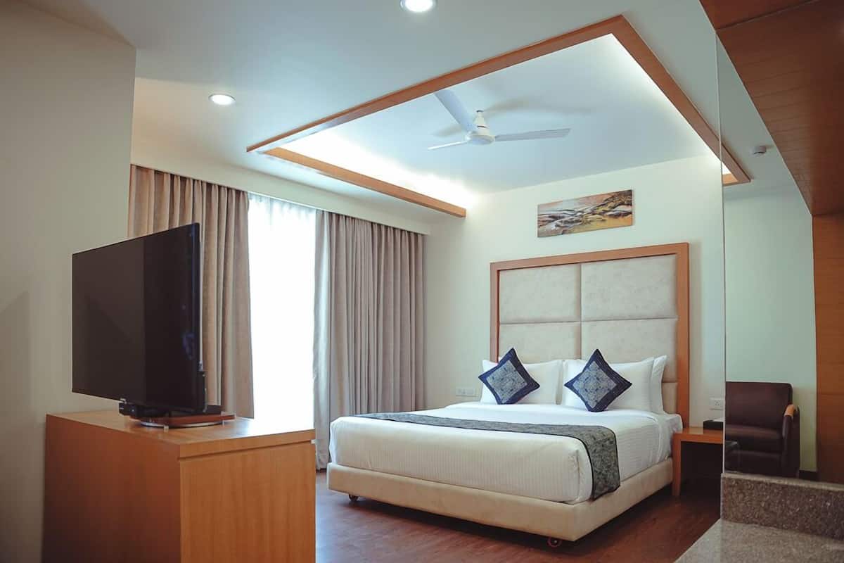 A hotel room with a large bed, a flat-screen TV on a wooden stand, a ceiling fan, and a painting on the wall.