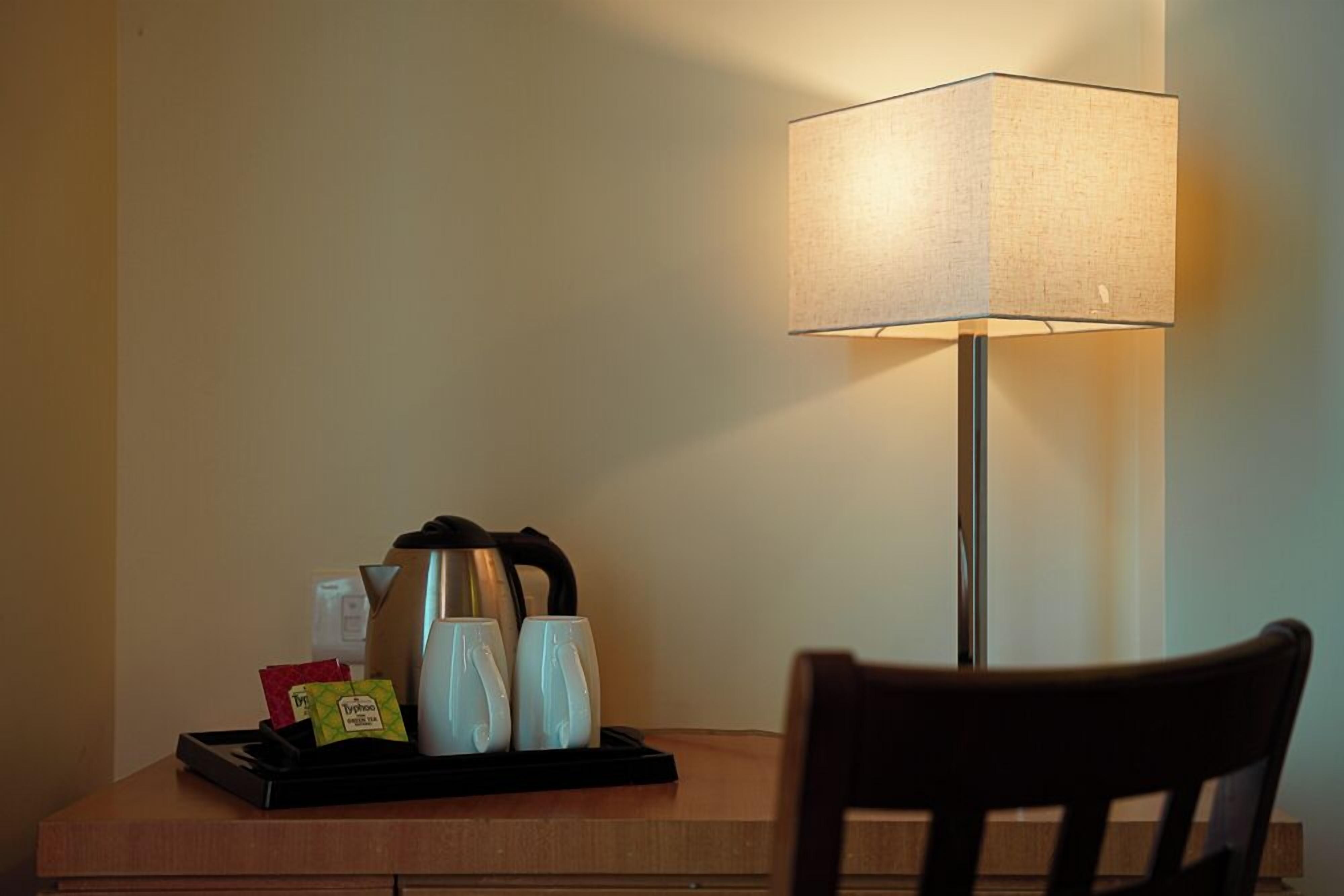 A hotel room with a desk, a tray with coffee cups and a kettle, and a lamp.