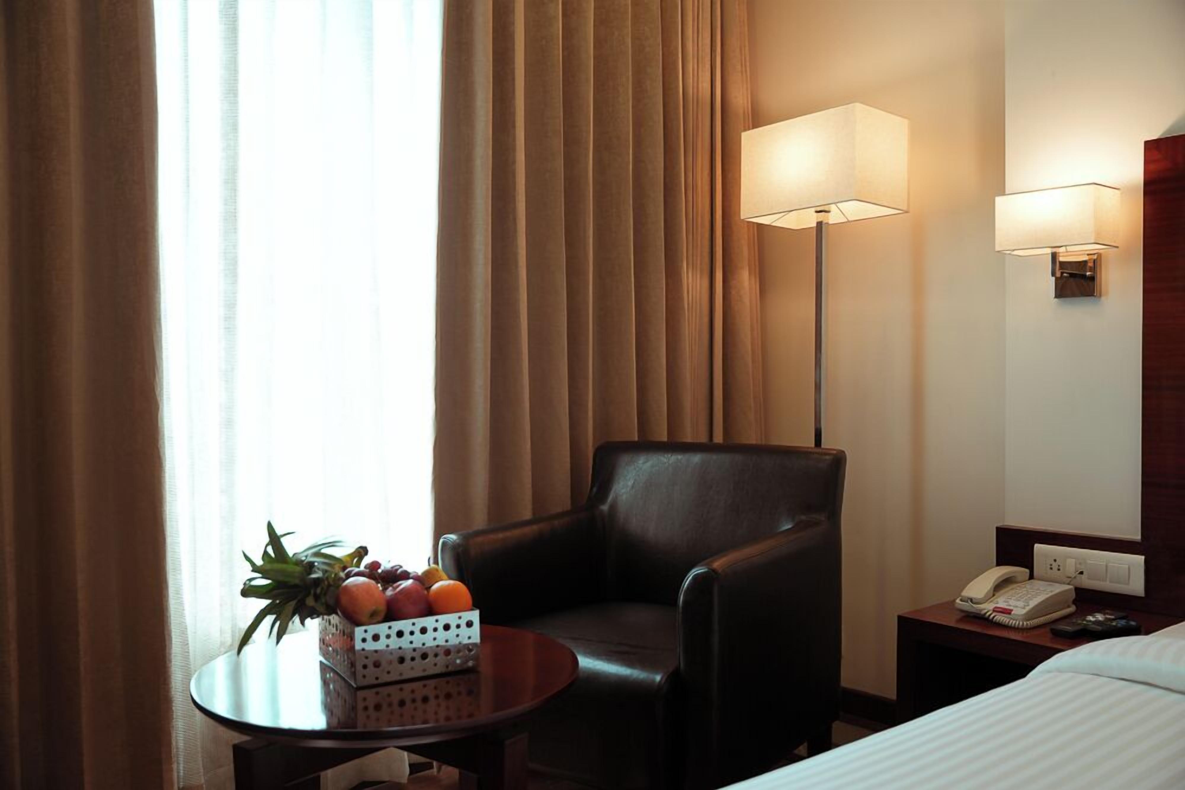 Une chambre d’hôtel avec un fauteuil en cuir, une petite table ronde sur laquelle se trouve un panier de fruits, une table de chevet avec un téléphone et une lampe.