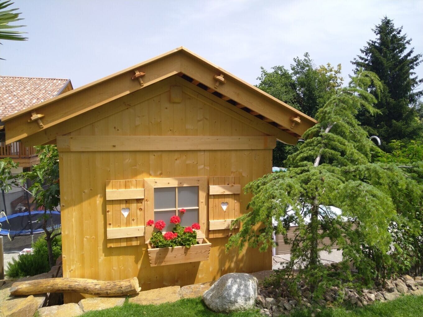 A wooden cabin with a pitched roof, two windows with heart-shaped cutouts, and a flower box with red flowers.