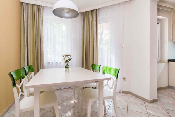 A dining area with a white table and chairs, a vase with flowers, and a kitchen in the background.