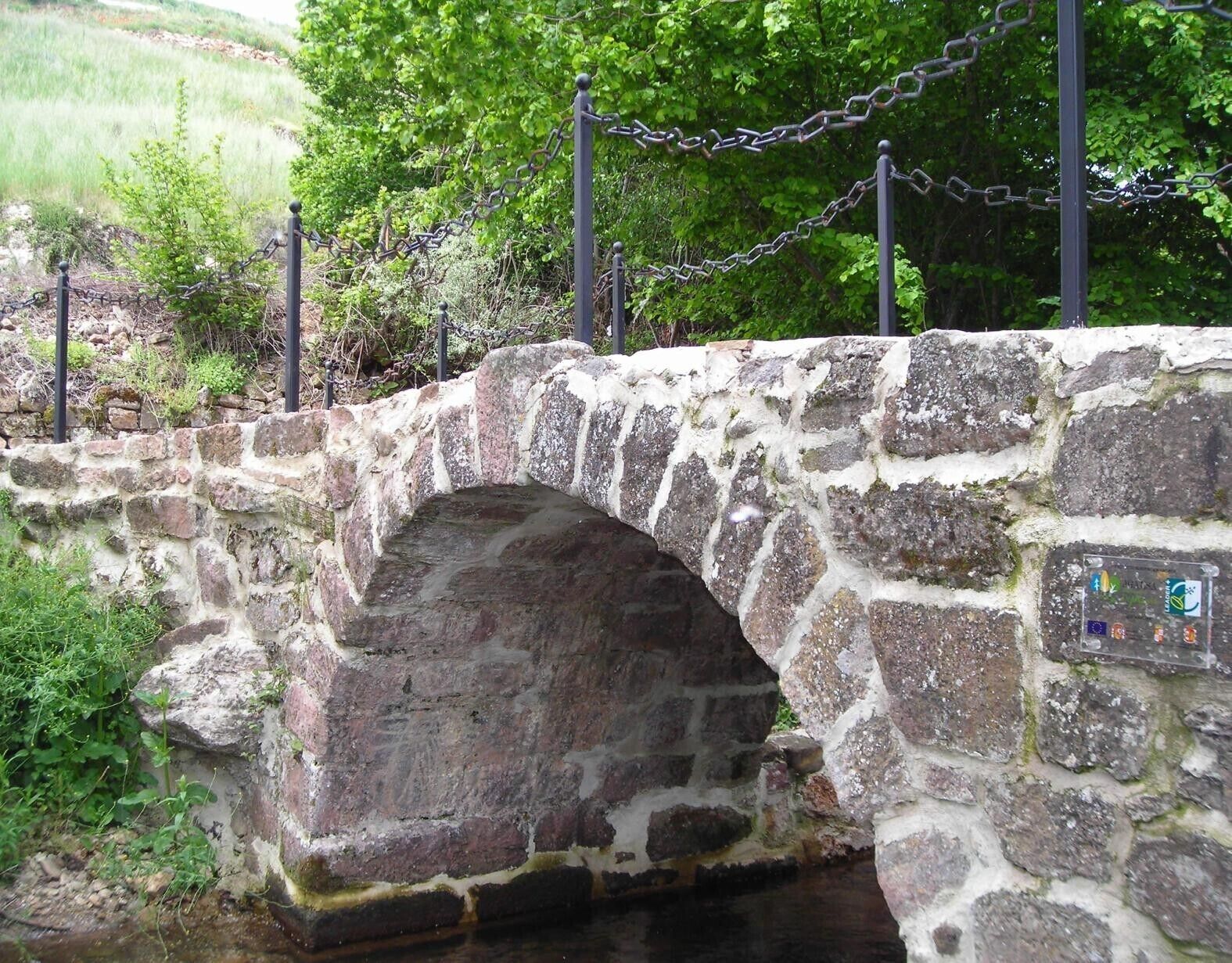 Un puente de piedra con una barrera de cadena metálica, rodeado de vegetación.