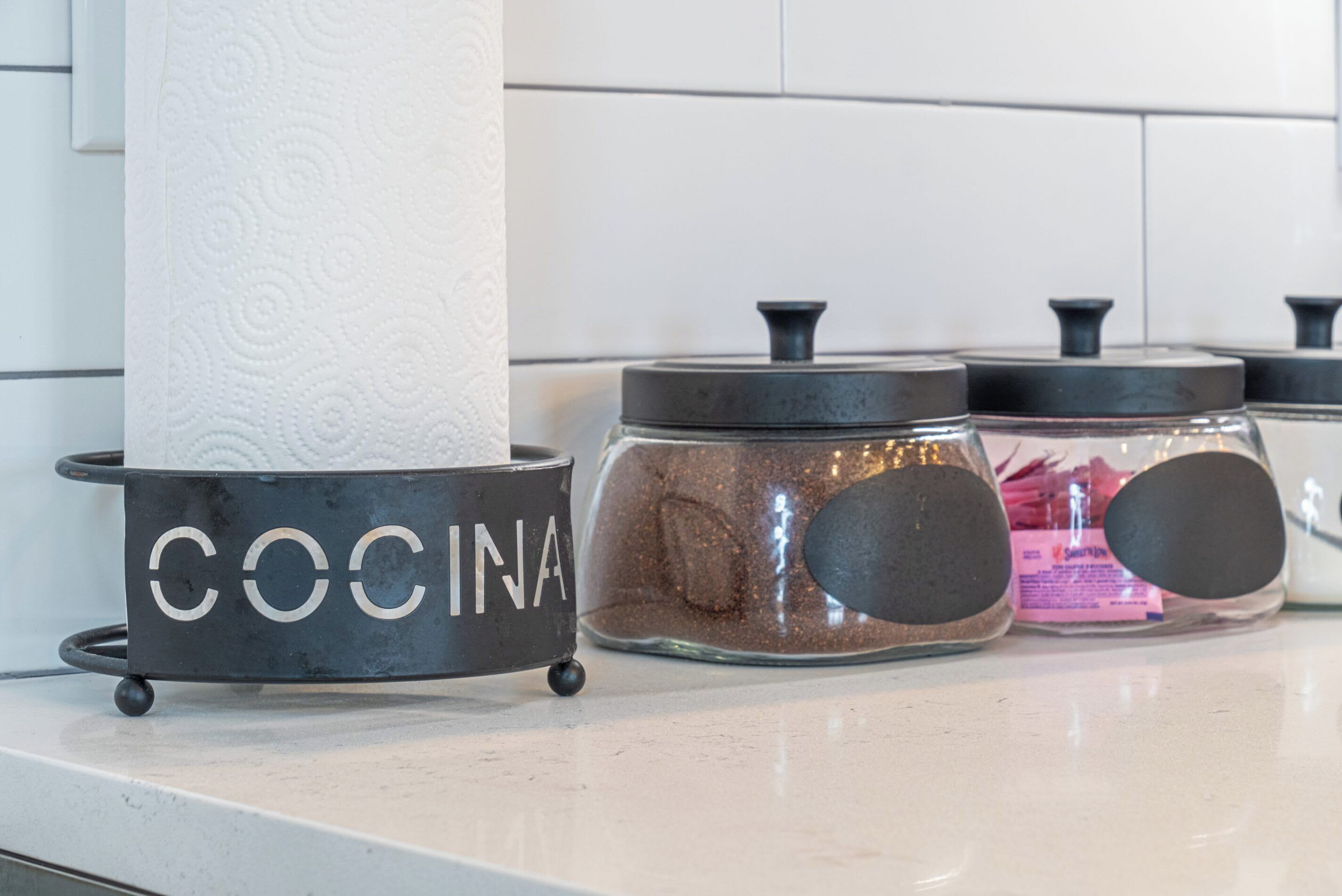 A kitchen counter with a roll of paper towels, a metal container labeled 'COCINA', and two glass jars with black lids.
