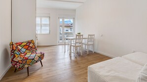 A room with a wooden floor, a white bed, a colorful chair, a dining table with chairs, and a view through a door.