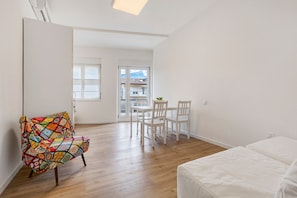 Une pièce avec un plancher en bois, un lit blanc, une chaise colorée, une table à manger avec des chaises, et une vue sur l’extérieur, visible à travers une porte.