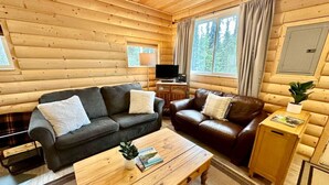 A cozy log cabin interior with a sofa, a leather couch, a coffee table, and a small kitchen area.