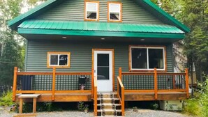 A two-story cabin with a green roof, a front door, and a small porch with a wooden railing.
