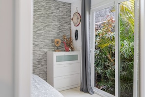 A modern bedroom with a bed, a dresser, a wall-mounted shelf with decorative items, and a view of a garden through a glass door.