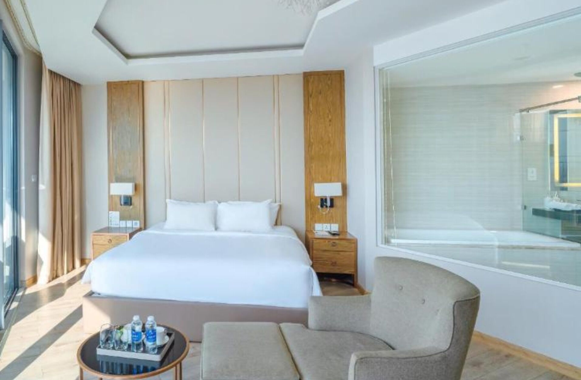 A hotel room with a large bed, two bedside tables, a chair, a small table with water bottles, and a large window.