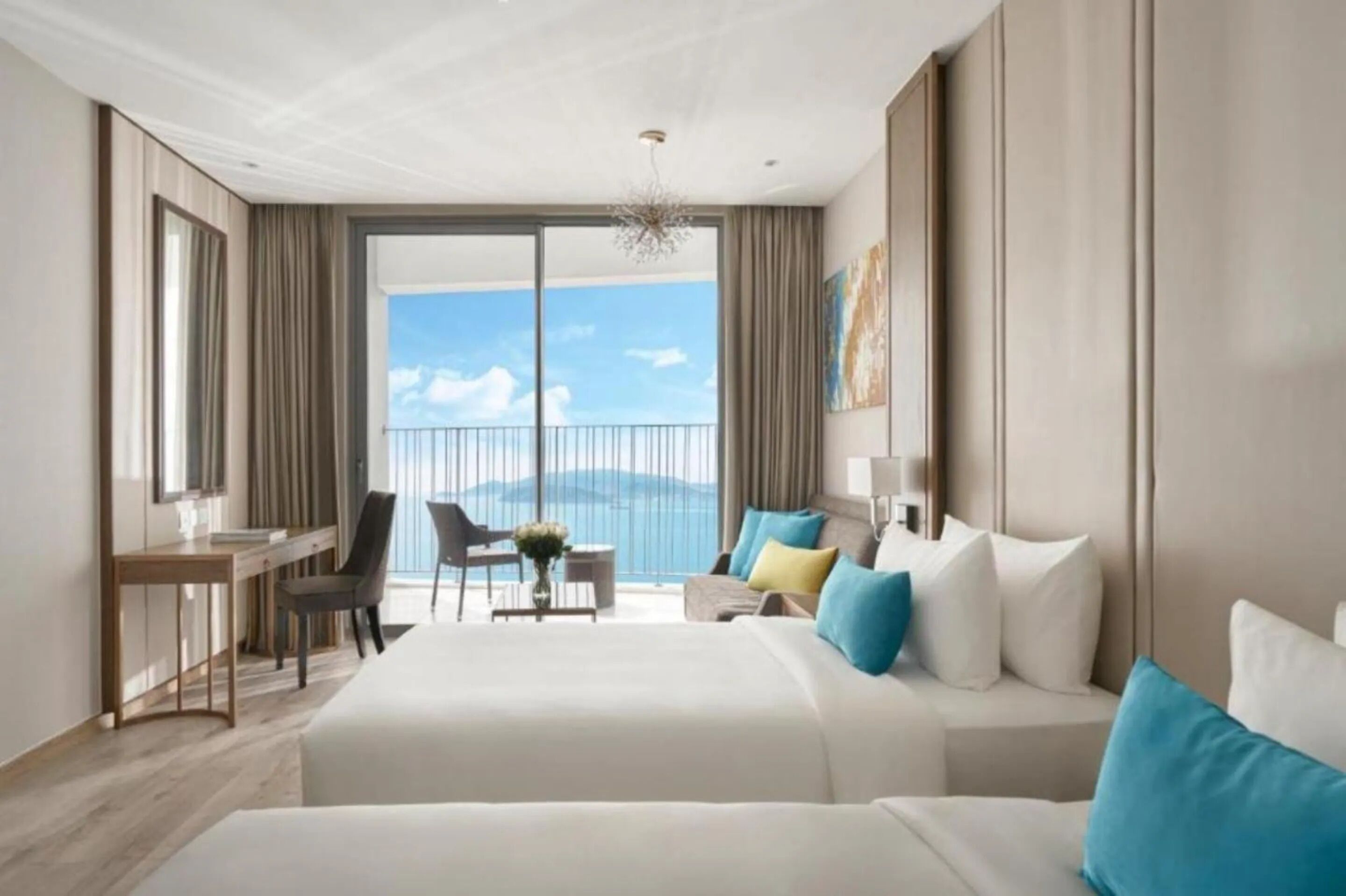 A modern hotel room with a large bed, a desk, and a chair. There is a balcony with a view of the sky and mountains.