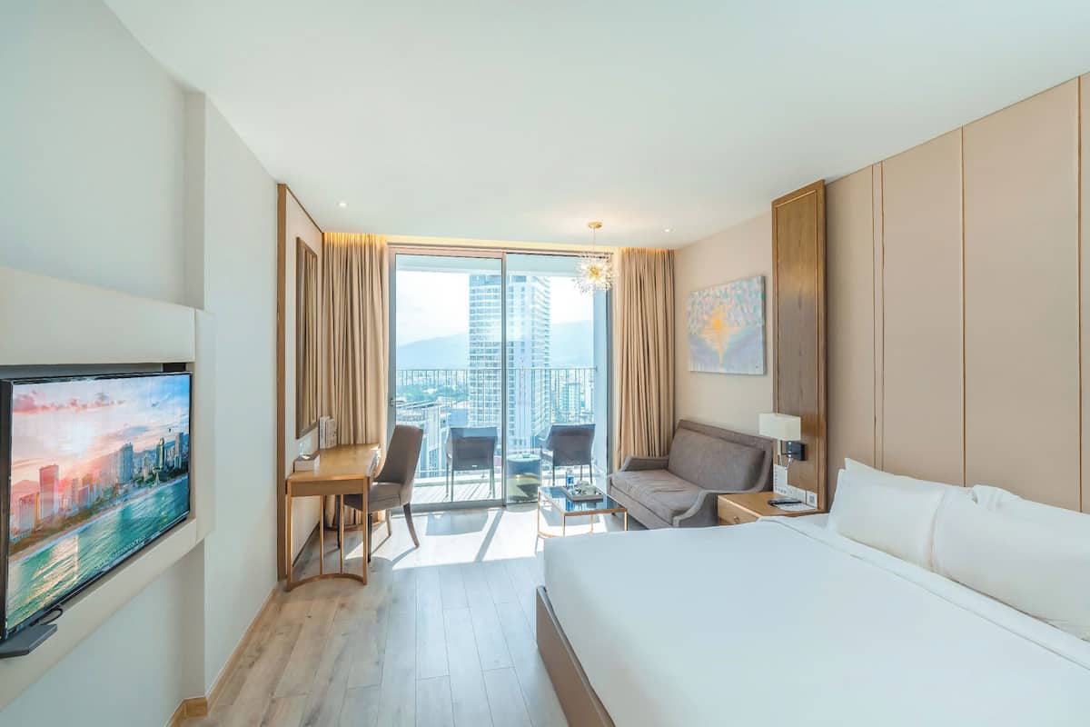 Een hotelkamer met een groot bed, een bureau met stoel, een bank, een televisie aan de muur en een balkon met uitzicht op de stad.