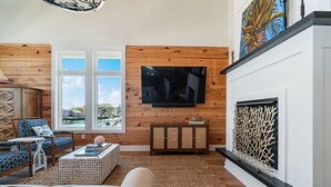 A living room with a fireplace, a television mounted on a wooden wall, and a view of the outdoors through large windows. - Sea Salt Cottage with private boat dock (Atlantic Beach)