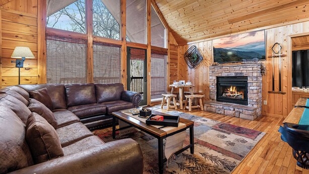 A cozy cabin interior with a stone fireplace, leather sofa, and large windows. - Near Dollywood*Easy Access Roads*Jacuzzi*Pool Table (SEVIERVILLE)