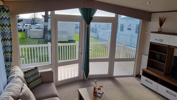 A living room with a large sliding glass door leading to a backyard with a white fence and a lawn. - Stunning Seaside Lodge Nr New Forest 2 bed (New Milton)