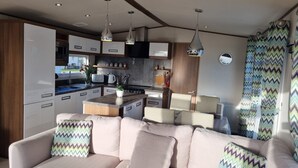 A modern kitchen with white cabinets, a central island, and pendant lighting. There is a beige sofa with patterned cushions in the living area. - Stunning Seaside Lodge Nr New Forest 2 bed (New Milton)