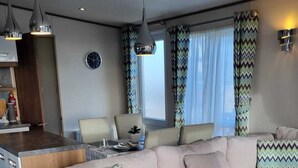 A modern living room with a beige sofa, a wooden coffee table, and a kitchen area with a dining table and chairs. - Stunning Seaside Lodge Nr New Forest 2 bed (New Milton)