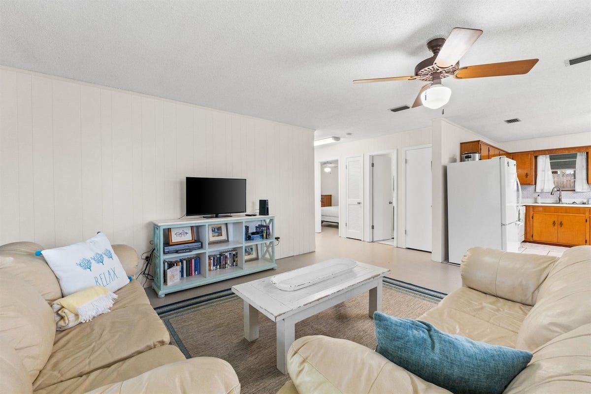 A living room with a sofa, a coffee table, and a television. There is a ceiling fan and a bookshelf with books.